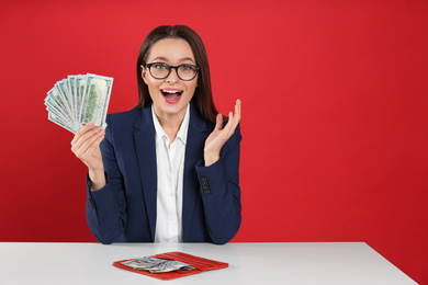 Emotional young woman with money and wallet at table on crimson background