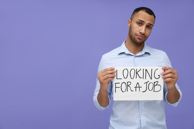 Photo of Young unemployed man holding sign with phrase Looking For A Job on violet background. Space for text
