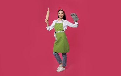 Young housewife in oven glove holding rolling pin on pink background