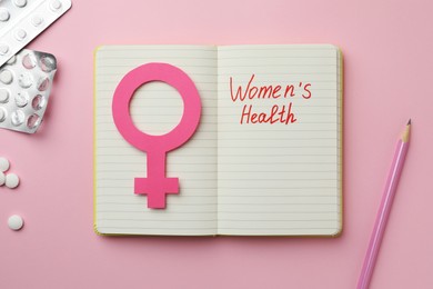 Notebook with female gender sign, pills and text Women's Health on pink background, flat lay