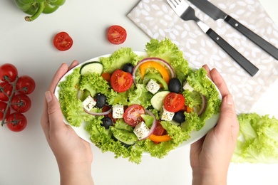 Woman holding plate of tasty fresh Greek salad over white table, top view