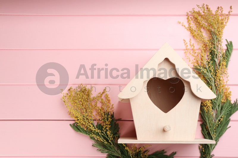 Beautiful bird house with heart shaped hole and mimosa flowers on pink wooden background, space for text. Spring flat lay composition