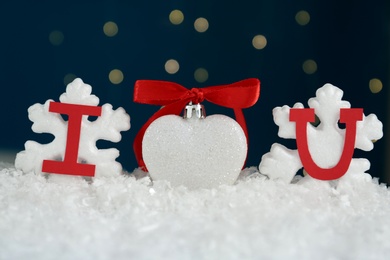 Phrase I Love You made of paper letters and festive heart shaped ornament on snow against blue background