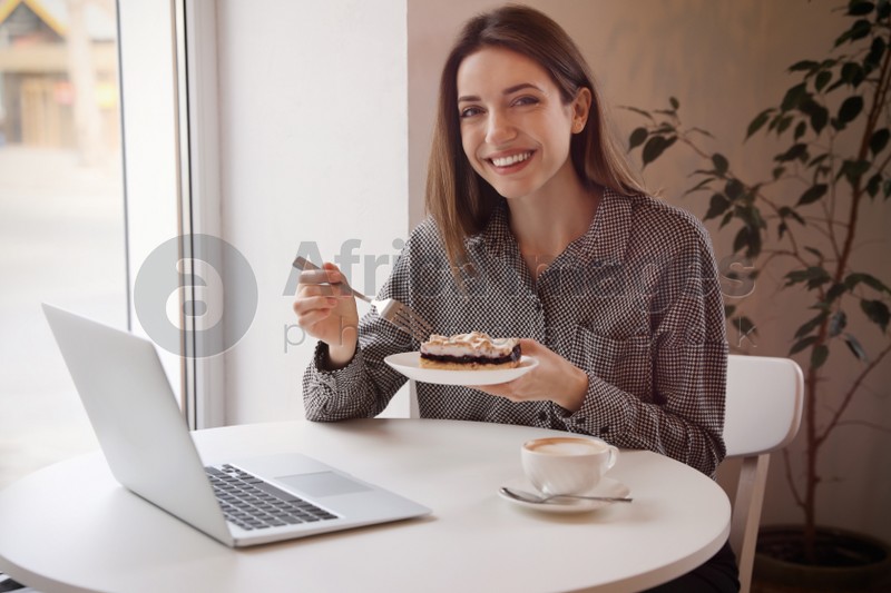 Young blogger with laptop eating dessert at table in cafe