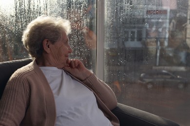 Elderly woman looking out of window on rainy day, space for text. Loneliness concept