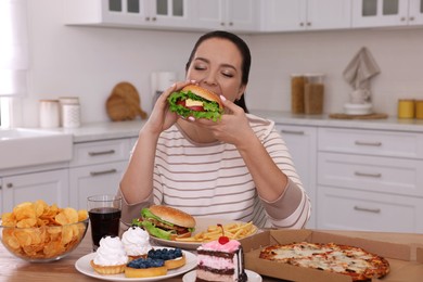 Photo of Overweight woman eating burger in kitchen at home