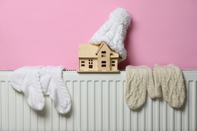 Modern radiator with knitted hat, socks, mittens and wooden house near pink wall indoors. Winter heating efficiency