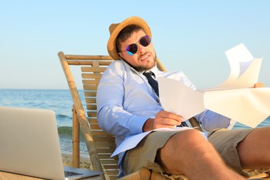 Photo of Busy man with papers talking by mobile phone on beach. Business trip