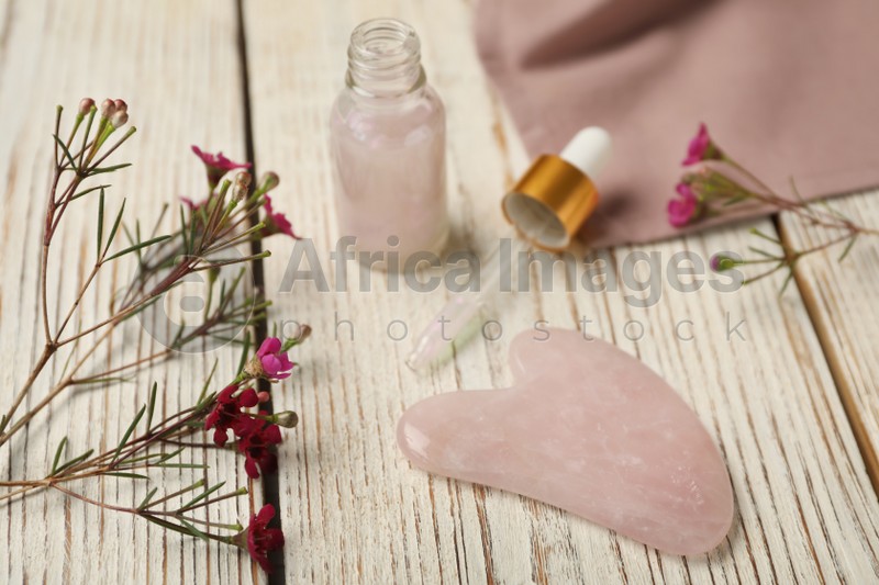 Photo of Rose quartz gua sha tool, skin care product and flowers on white wooden table
