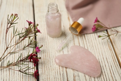 Photo of Rose quartz gua sha tool, skin care product and flowers on white wooden table