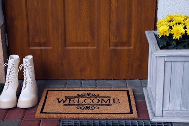 Photo of Doormat with word Welcome, stylish boots and beautiful flowers on floor near entrance