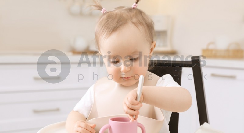 Cute little baby with spoon and cup in high chair indoors