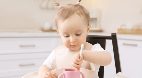 Cute little baby with spoon and cup in high chair indoors