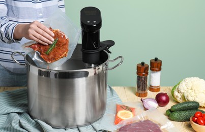 Woman putting vacuum packed meat into pot with sous vide cooker at wooden table, closeup. Thermal immersion circulator