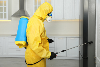 Pest control worker in protective suit spraying insecticide near refrigerator indoors