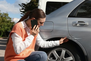 Stressed woman talking on phone near car with scratch outdoors