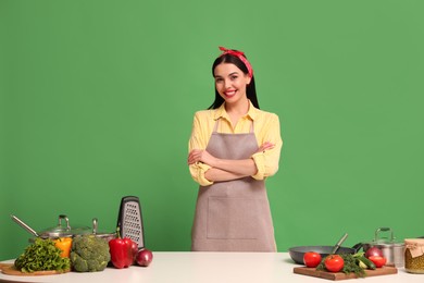Young housewife at white table with vegetables and different utensils on green background