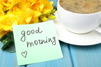 Photo of Cup of aromatic coffee, beautiful yellow freesias and Good Morning note on light blue wooden table, closeup