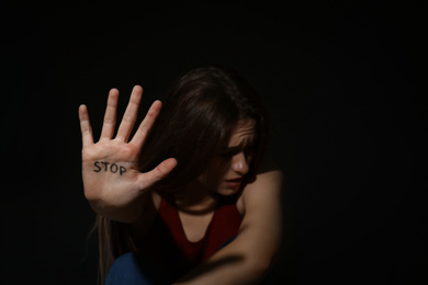 Abused young woman showing palm with word STOP against black background, focus on hand. Domestic violence concept