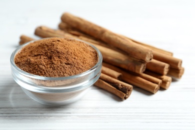 Aromatic cinnamon powder in bowl near sticks on white table, closeup