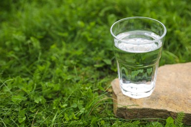 Glass of fresh water on stone in green grass outdoors. Space for text