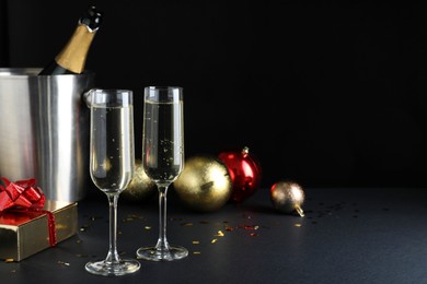 Happy New Year! Glasses with sparkling wine, bottle in bucket and festive decor on table against black background, space for text