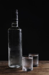 Bottle of vodka and shot glasses on wooden table against black background. Space for text