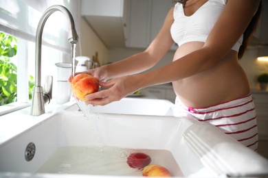 Photo of Young pregnant woman washing fresh sweet peach in kitchen, closeup. Taking care of baby health