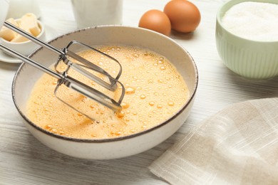 Beating eggs in bowl with mixer at white wooden table, closeup