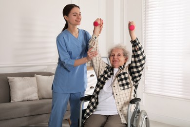 Senior woman in wheelchair doing physical exercise and young caregiver helping her indoors. Home health care service