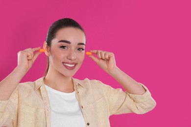 Young woman inserting foam ear plugs on pink background