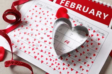 Calendar with marked Valentine's Day, red ribbon and heart shaped cookie cutter on grey table, closeup