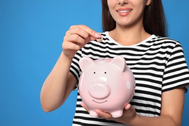 Young woman putting coin into piggy bank on light blue background, closeup