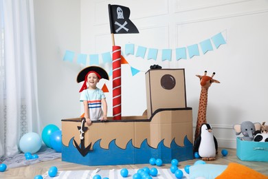 Cute little boy playing with pirate cardboard ship and toys at home. Child's room interior