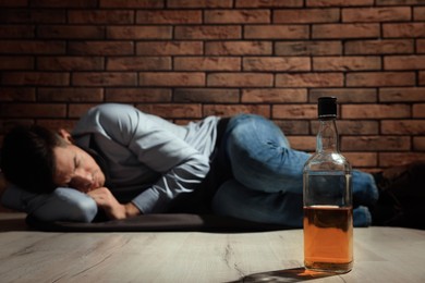 Addicted man lying near red brick wall, focus on bottle of alcoholic drink