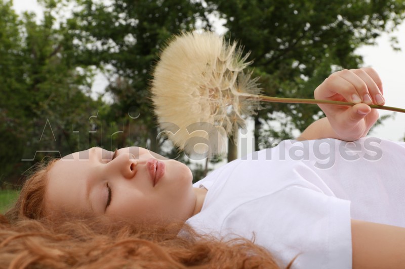 Photo of Cute girl with beautiful red hair blowing large dandelion while lying on green grass in park. Allergy free concept