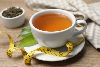 Cup of herbal diet tea and measuring tape on wooden table, closeup. Weight loss concept