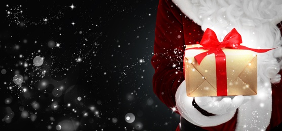 Santa Claus holding gift box on black background with snowflakes, bokeh effect. Space for text