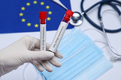 Doctor holding sample tubes with labels Coronavirus Blood Test above medical items and European Union flag, closeup