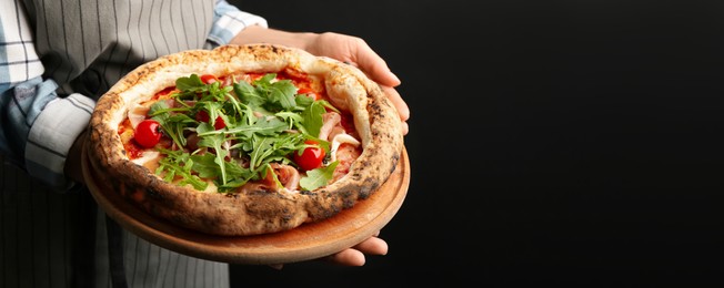Woman holding tasty pizza with meat and arugula on black background, closeup view with space for text. Banner design