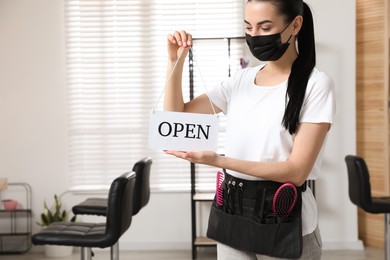 Professional stylist with protective mask holding Open sign in salon, space for text. Hairdressing services during Coronavirus quarantine