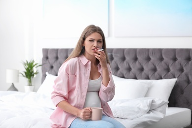 Young pregnant woman with cup of coffee smoking cigarette in bedroom. Harm to unborn baby