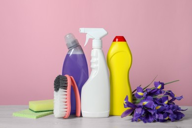 Photo of Different cleaning supplies and beautiful spring flowers on white wooden table against light pink background