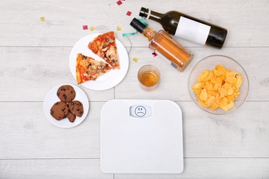 Digital scales, junk food and alcohol on wooden floor after party, top view