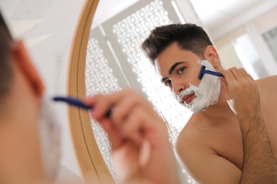 Handsome young man shaving with razor near mirror in bathroom