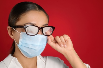 Woman wiping foggy glasses caused by wearing medical mask on red background, closeup