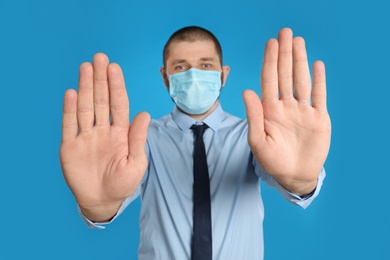 Man in protective mask showing stop gesture on light blue background. Prevent spreading of coronavirus