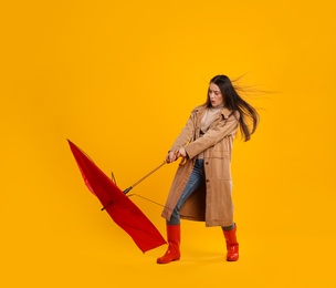 Emotional woman with umbrella caught in gust of wind on yellow background