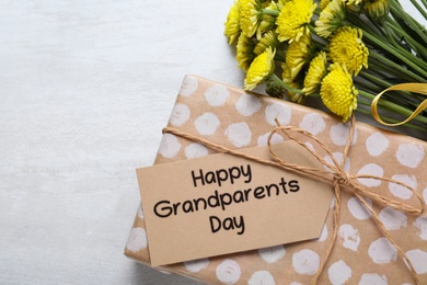 Beautiful yellow flowers, gift box and tag with phrase Happy Grandparents Day on light background, flat lay