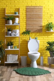 Stylish bathroom with toilet bowl and green plants near yellow brick wall. Interior design